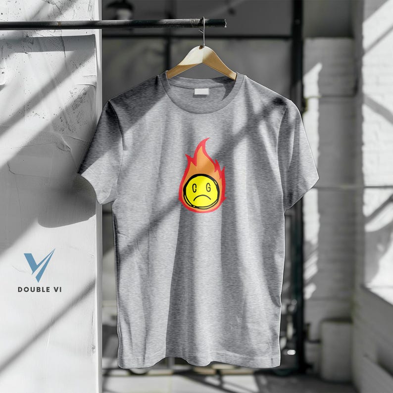 May include: A heather gray t-shirt with a graphic of a sad yellow face engulfed in red and orange flames. The letters "C G" are inside the yellow face. The shirt is hanging on a wooden hanger.