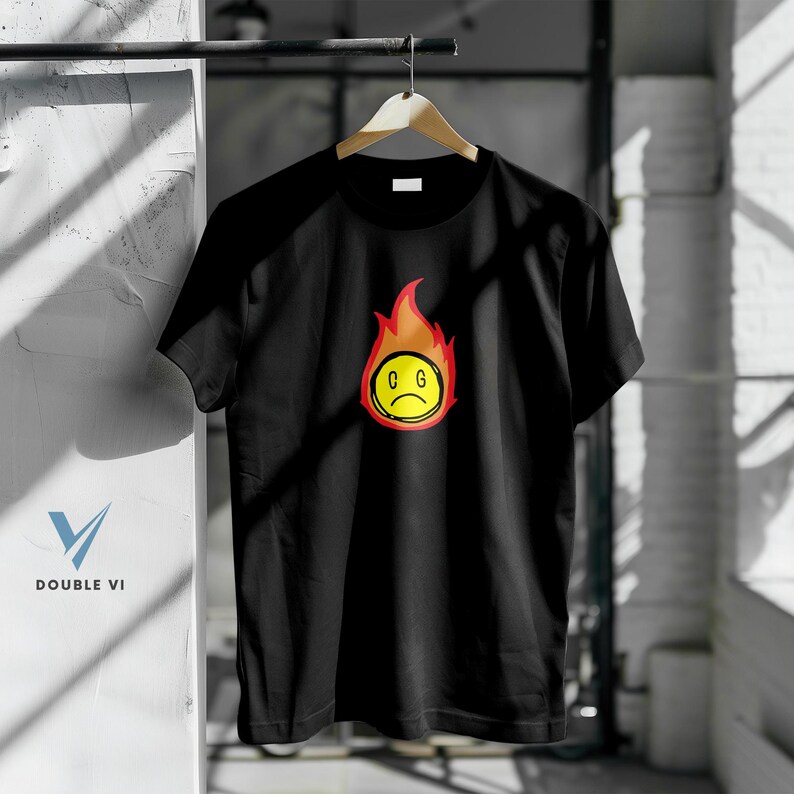May include: Black t-shirt with a graphic of a yellow, sad-faced emoji surrounded by red and orange flames. The emoji has the letters "C G" inside. The shirt is hanging on a wooden hanger.
