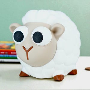 May include: A whimsical sheep-shaped piggy bank. The bank features a white, fluffy body, a tan face with large, expressive eyes, and brown hooves. The coin slot is on the top. A fun and functional item for kids.