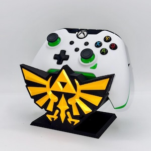 May include: A white video game controller with green buttons and a black and gold Triforce stand. The stand is a 3D printed design of the Triforce symbol from the Legend of Zelda video game series.