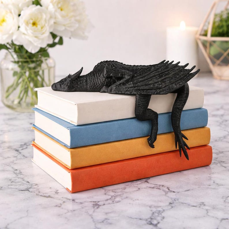 May include: A black dragon figurine resting on a stack of four books. The dragon has textured scales, folded wings, and a long tail draped over the side. The books are white, blue, yellow, and orange, arranged in a vertical column on a surface.