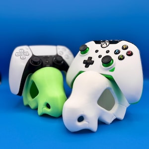 May include: Two white video game controllers with green and black accents. The controllers are resting on top of two white and green skull-shaped objects.