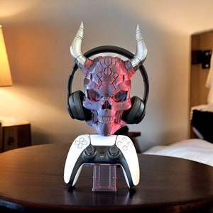 May include: A red and black skull-shaped headset stand with horns, holding a white wireless gaming controller and black headphones.