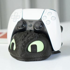 Könnte beinhalten: Ein weißer PlayStation 5-Controller liegt auf einem schwarzen Controller-Ständer im Drachen-Design. Der Drache hat leuchtend grüne Augen. Der Ständer ist so konzipiert, dass er den Controller sicher hält.