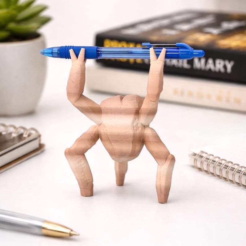May include: A unique desk accessory featuring a wooden sculpture of a figure with arms raised, holding a blue pen. The sculpture has a light brown color and is positioned on a white surface, with a spiral notebook and a pen in the background.