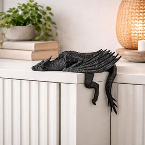 May include: A black dragon figurine with textured scales and folded wings, positioned to hang over the edge of a white surface. The creature features a long tail, sharp claws, and a detailed head, crafted in a solid matte black finish for display.