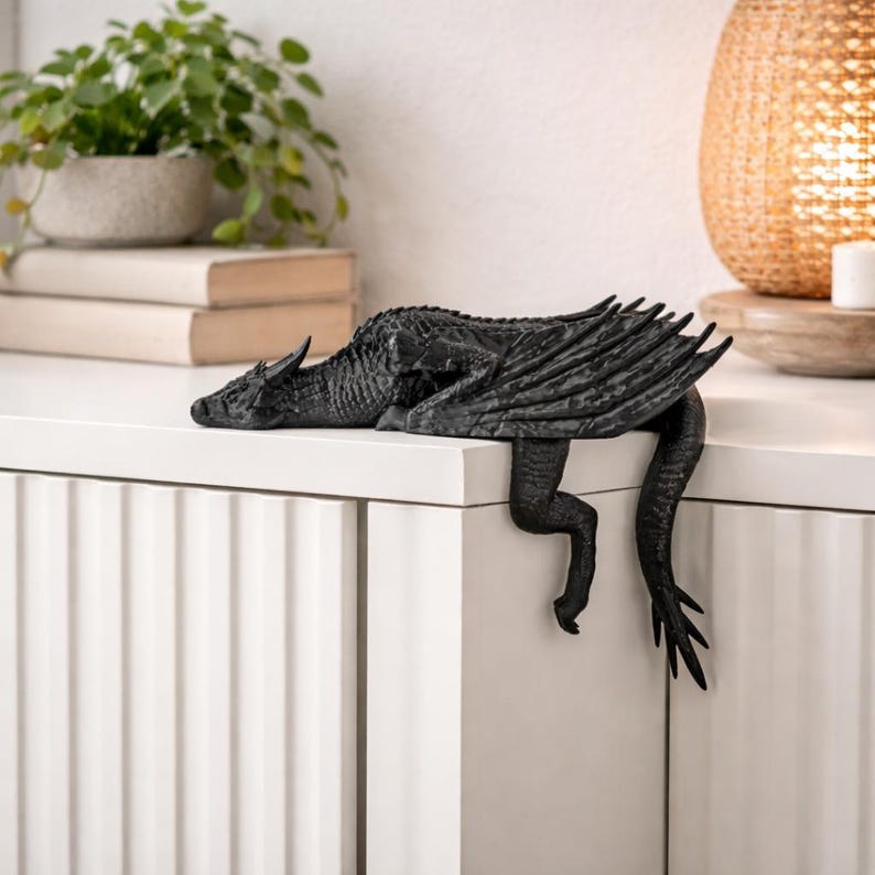 May include: A black dragon figurine with textured scales and folded wings, positioned to hang over the edge of a white surface. The creature features a long tail, sharp claws, and a detailed head, crafted in a solid matte black finish for display.