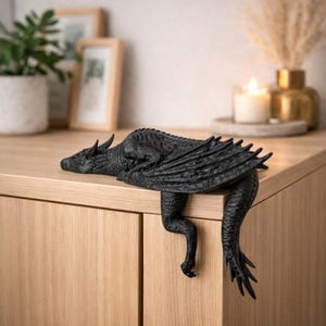 May include: A matte black dragon figurine designed to perch on the edge of a surface. The sculpture features detailed scales, folded wings, and a long tail that hangs down over the side. The dragon is positioned in a resting pose with its head.