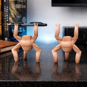May include: Two wooden pen holders shaped like figures, one holding a pen aloft. The figures are light brown with four legs and arms raised. The pen is black. The figures are on a dark, reflective surface.
