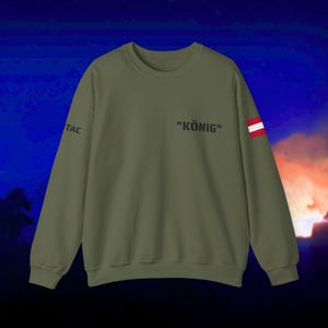 May include: An olive green crewneck sweatshirt featuring the text "KÖNIG" in black on the upper chest. The right sleeve displays the text "TAC" in black, while the left sleeve features a small rectangular patch with the Austrian flag design.