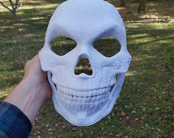 May include: White skull mask with intricate floral and swirl designs. The mask is made of a smooth, white material and has eye and mouth openings.