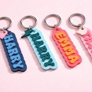 May include: Four keyrings with names: "HARRY", "EMMA", and "TAYLOR". Each keyring has a silver ring and a decorative charm. The "HARRY" keyring is navy blue with light blue letters and a pink heart charm.