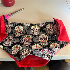Può includere: Una borsa di tessuto nero e rosso con un motivo di teschio di zucchero. La borsa è cucita con filo rosso e ha una fodera rossa.
