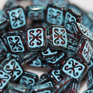May include: A close-up of a string of square-shaped glass beads. The beads are a deep blue color with a red-brown glaze and a white design. The design is a stylized floral pattern.