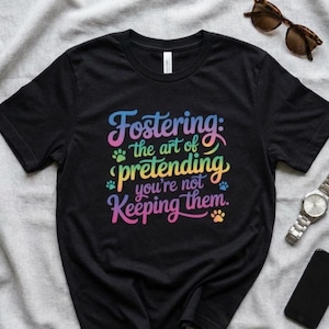 Peut inclure: T-shirt noir avec le texte "Fostering: the art of pretending you're not keeping them" dans une police colorée en arc-en-ciel. Le t-shirt comporte également des empreintes de pattes. Des lunettes de soleil, une montre et un téléphone sont également visibles.