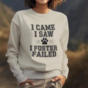 Peut inclure: Un sweat-shirt gris clair avec le texte "I CAME I SAW I FOSTER FAILED" en lettres noires. Un motif de patte de chien est centré entre "SAW" et "I". La personne porte un pantalon de survêtement gris clair.
