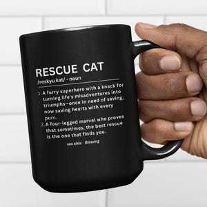 May include: Black ceramic mug with the text "RESCUE CAT" and a definition. The definition describes a rescue cat as a furry superhero. The mug has a large handle and is held by a person.