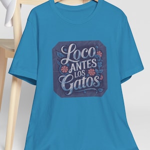 Op de afbeelding: Een turquoise blauw t-shirt met een grafische print van een vierkant met de tekst "Loco Antes Los Gatos" in een gestileerd lettertype met bloemmotieven.