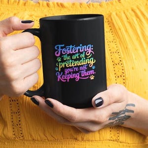 Peut inclure: Une tasse en céramique noire avec une grande anse. La tasse présente un texte coloré qui dit "Fostering: the art of pretending you're not keeping them." Des empreintes de pattes sont incluses dans le design.