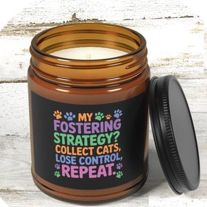 Pode incluir: Uma vela de pote de vidro castanho com uma tampa preta. A vela tem um rótulo preto com texto colorido que diz "MY FOSTERING STRATEGY? COLLECT CATS, LOSE CONTROL, REPEAT." As pegadas são usadas como elementos de design.