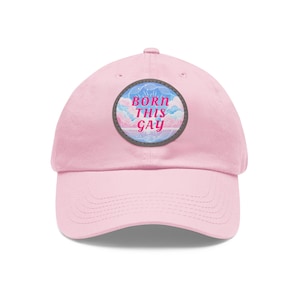 May include: A light pink baseball cap with a curved brim. The cap features a circular patch with the words "BORN THIS GAY" in pink and blue, set against a cloudy sky design. The hat is a classic, casual accessory.
