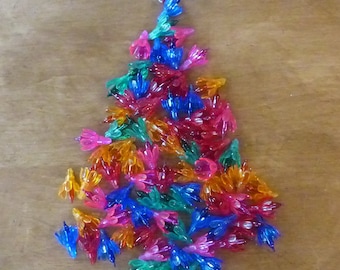 May include: A colorful Christmas tree decoration made of translucent plastic flowers in shades of pink, orange, blue, and green. The tree is topped with a white flower. The background is a light brown wooden surface.