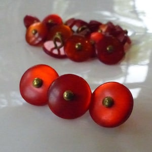 May include: A collection of red, translucent buttons with brass-colored centers. The buttons are round and have a slightly domed shape. Some buttons are in focus, while others are blurred in the background.