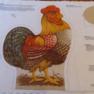May include: A fabric panel featuring a colorful rooster design. The rooster has a red comb and wattle, with brown, orange, and white feathers. The design is cut out, ready for crafting. Instructions and a cardboard oval are also visible.