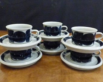 May include: Set of six white ceramic coffee cups and saucers with a blue floral pattern. The cups and saucers are stacked on a wooden table.
