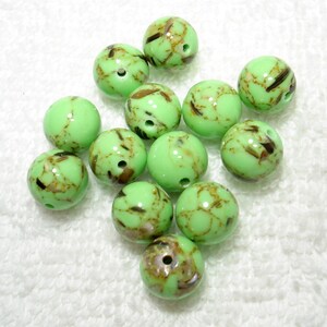 May include: A collection of round, light green beads with brown and tan marbling. Each bead has a small hole for stringing. The beads are scattered on a white, textured surface. These beads are suitable for jewelry making and crafts.