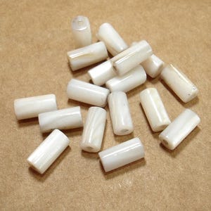 May include: A collection of white, cylindrical beads with a glossy finish. The beads are scattered on a light brown surface. Each bead has a hole running through the center, and they vary slightly in size and shape.