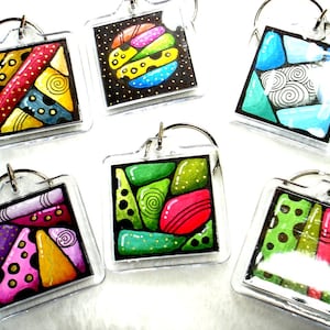 May include: A collection of six square acrylic keychains with colorful abstract designs. Each keychain features a different pattern of shapes and colors, including red, yellow, blue, green, and black. The keychains have silver-colored key rings.