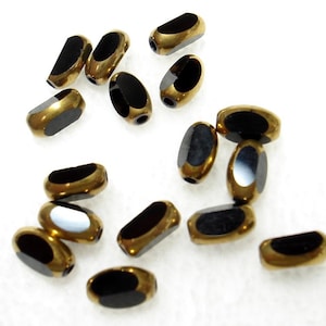 May include: A collection of oval-shaped beads. Each bead has a black, glossy center and a metallic gold-colored border. The beads are scattered on a white surface, showcasing their design and texture. Ideal for jewelry making.