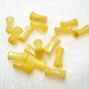 May include: A collection of yellow, hourglass-shaped beads scattered on a white surface. The beads have a glossy finish and a slightly translucent appearance, with variations in color intensity.