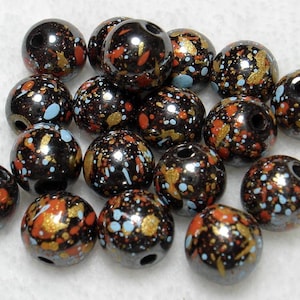 May include: A collection of round, dark gray beads featuring a glossy finish and a speckled pattern. Each bead is decorated with small, irregular paint splatters in vibrant shades of orange, metallic gold, and light blue across the surface.