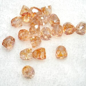 May include: A collection of small, faceted, translucent beads in a warm peach and amber hue. Each bead features a multi-faceted surface that reflects light, creating a sparkling effect. The beads are shaped like rounded cones with a central hole.