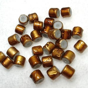 May include: A collection of small, barrel-shaped beads. The beads have a metallic bronze finish with gold accents and a light gray interior. These beads are suitable for jewelry making and crafting projects.