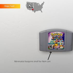 May include: A gray wall-mounted shelf designed to hold a Nintendo 64 game cartridge. A gray Nintendo 64 game cartridge is on the shelf. The game cartridge has a label with the text "Pokémon Puzzle League" and a picture of Pikachu and other Pokémon characters. The shelf is labeled "Minimalist footprint shelf for N64 cart".