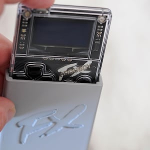 May include: A handheld gaming device, the Arduboy, is partially inserted into a light gray case. The device has a small screen and buttons. The case features the embossed letters "FX".