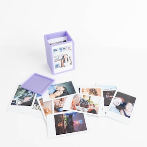 May include: A light purple square storage box with a matching lid, designed to hold small square photographs. The box is open, revealing a stack of various candid color photographs inside, while several other similar photographs are scattered around.