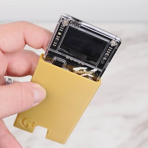 May include: A portable electronic gaming device with a clear transparent top section and a solid mustard yellow plastic base. The device features a small screen, visible internal circuitry, and the word ARDUBOY printed in white on the front surface.