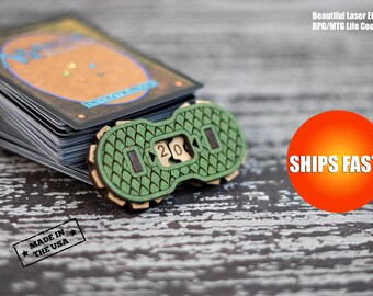 May include: Green and brown wooden life counter with a number 20 on it. The counter is made in the USA and ships fast.  The counter is sitting on top of a stack of Magic: The Gathering cards.
