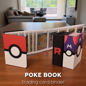 Könnte beinhalten: Auf einem Holztisch sind drei Sammelkartenordner zu sehen. Die Ordner sind als "Poke Book" gestaltet und haben rote, schwarze und weiße Farbtöne. Verschiedene Sammelkarten sind im Inneren sichtbar. Der Text "Trading card binder" steht darunter.