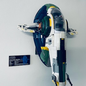 Puede incluir: Un modelo Lego de la nave espacial de Star Wars, Slave I, en blanco, verde, azul y amarillo. La nave está montada en una pared blanca. A la izquierda de la nave hay una placa negra con el texto "Star Wars Jango Fett's Starship".
