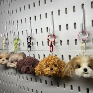 May include: Five small, fluffy dog-shaped keychains hanging from hooks on a pegboard wall. The keychains are made of yarn and have different coloured fur, including brown, white, and blonde. Each keychain has a small, black nose and eyes.