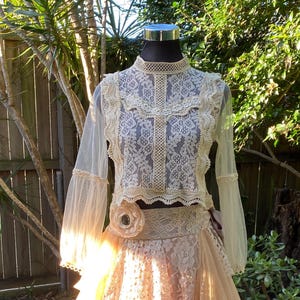 May include: A cream and peach two-piece outfit displayed on a mannequin. The top is a lace crop top with long, sheer sleeves. The skirt is a layered design in peach and cream tones, creating a romantic, bohemian aesthetic, perfect for a special occasion.