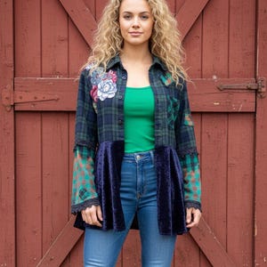 May include: A woman in a green top, blue jeans, and brown cowboy boots. She is wearing a plaid shirt with floral embroidery and a navy blue velvet peplum. The background is a red wooden door.