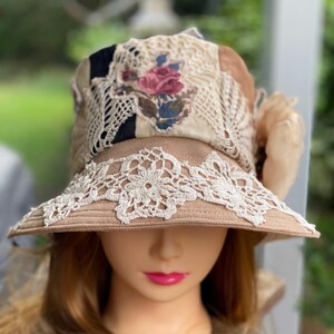 May include: A tan sun hat with a wide brim, decorated with cream lace and a floral patch. The hat has a patchwork design with black and tan fabric and a cream lace overlay. A peach fabric flower is attached to the side.