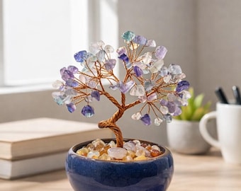 May include: A decorative tree sculpture with a copper wire trunk and branches, adorned with colorful gemstone leaves. The tree sits in a small, blue ceramic pot filled with pebbles. Two small stones are next to the pot. The sculpture is on a wooden surface.