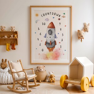 May include: A white wooden playhouse with yellow wheels, a rocking chair, a beanbag chair, and a framed print of a rocket ship with a countdown from 10 to 0. The print has a watercolor background with stars and planets. There are also stuffed animals on a shelf and hanging from hooks.